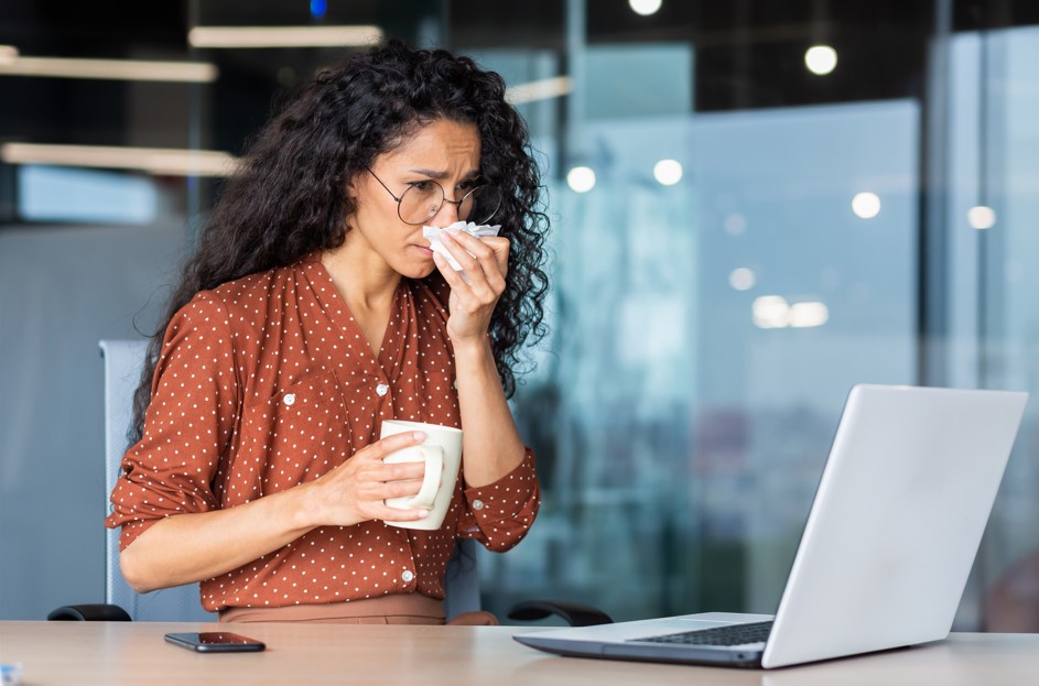 Alergias em ambientes de trabalho: como a limpeza pode minimizar? Mulher sentada à sua mesa de trabalho, claramente desconfortável devido a uma possível alergia ou resfriado. Ela está assoando o nariz e segura uma caneca, possivelmente com chá quente, indicativo de que está tentando aliviar seus sintomas. A cena destaca a importância de abordar alergias em ambientes de trabalho.